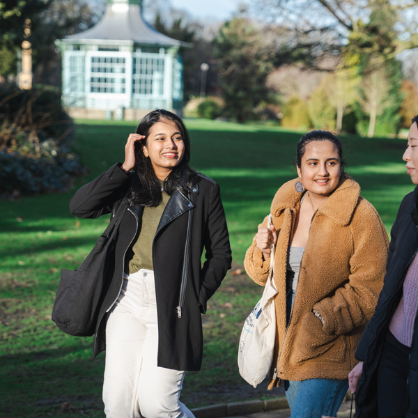 Three people walking together along a paved path in a park with green grass and trees. They are dressed in warm, casual clothing, including coats and jackets, and carrying bags. In the background, there is a light blue gazebo surrounded by greenery, and the scene is lit by natural daylight.