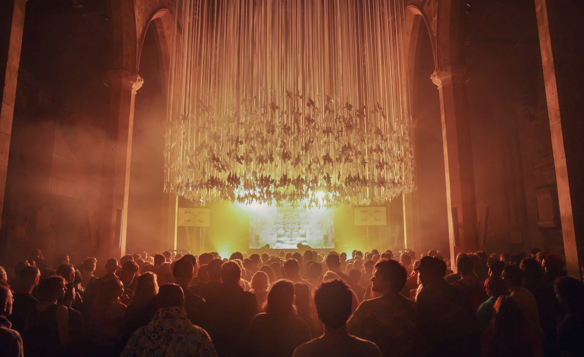 A large crowd gathered inside a grand hall with tall stone arches, facing a brightly lit stage. Above the audience hangs an elaborate installation of long, vertical ribbons or strands, illuminated by warm golden light that fills the space with a dramatic glow. The atmosphere suggests a concert or immersive art event in a historic architectural setting.