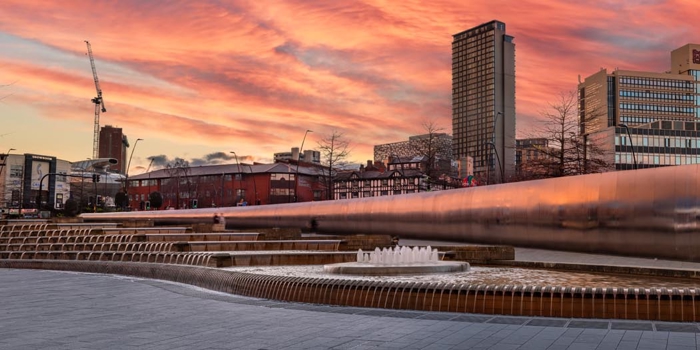 Urban plaza with cascading water features and fountains in front of a long reflective wall. Modern high-rise buildings and older brick structures are visible in the background, along with construction cranes. The sky is filled with dramatic orange and pink clouds during sunset, creating a vibrant backdrop.