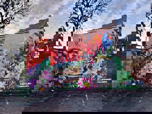 Large colorful mural on a brick wall depicting a vibrant scene with flowers, birds, a snail shell, and architectural elements under a twilight sky. A street sign reading ‘London Road’ is visible at the bottom right, and a tree partially frames the mural on the left.