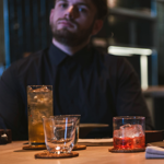 A wooden dining table in a modern restaurant setting with drinks placed on coasters. There is a tall glass of iced drink, a short glass with a pink cocktail and ice, and an empty tumbler. A folded napkin and cutlery are also visible, along with a small dish of food at the edge of the frame. The background shows a softly lit interior with sleek, contemporary design elements.