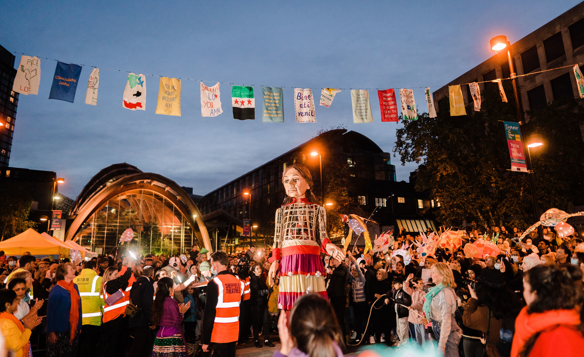 Outdoor evening street festival with a large crowd gathered around performers. Colorful flags hang overhead, and event staff in orange vests are visible. A tall figure in a decorative costume stands in the centre, with illuminated buildings and a glass arch structure in the background.