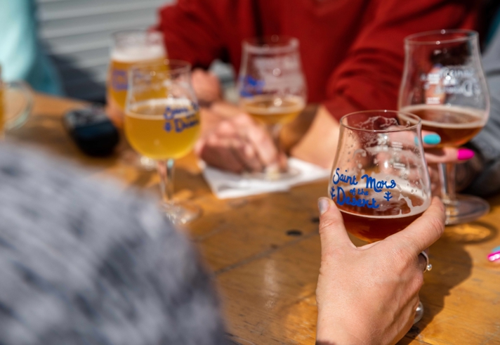 Close-up of several people sitting around a wooden table with multiple glasses of craft beer. One hand in the foreground holds a glass labeled ‘Saint Mars of the Desert.’ Other glasses with similar labels and varying shades of beer are visible, along with a notepad and pen on the table.