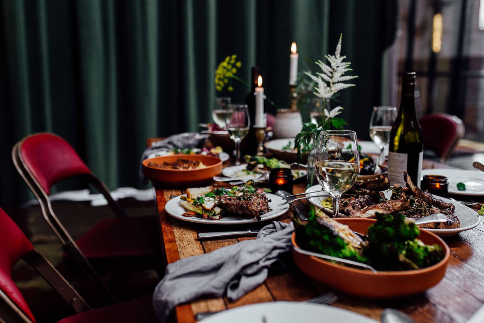 A table at The Mowbray full of food and wine. Elegant dining table set for a meal with plates of grilled meat, vegetables, and side dishes. The table features wine glasses, a bottle of wine, candles, and decorative greenery. Red chairs surround the wooden table, and a dark green curtain hangs in the background, creating a cozy, intimate atmosphere.