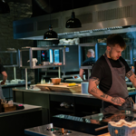 Chefs working in a modern open kitchen with stainless steel counters and cooking stations. One chef in the foreground, wearing a grey apron, is preparing food near an induction hob with small bowls and utensils. The background shows other chefs at work, shelves with plates, and a large oven. Black pendant lights hang from the ceiling, and the setting has an industrial, contemporary design.
