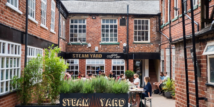 Outdoor courtyard of Steam Yard café with people sitting at tables, surrounded by red‑brick buildings and greenery.