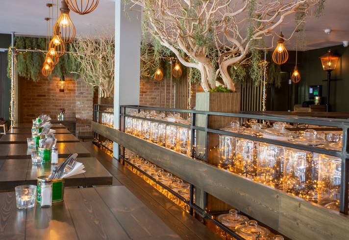 A stylish restaurant interior with long wooden tables, warm pendant lights, decorative trees in large planters and shelves of glass jars lit with fairy lights, creating a soft, ambient glow.
