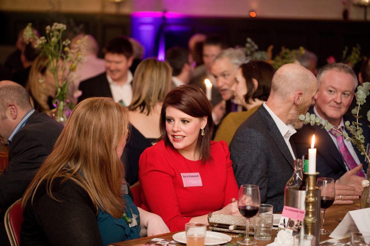 A formal dinner event with guests seated at a long wooden table decorated with tall floral arrangements and lit candles. The table is set with wine glasses, bottles of wine, and plates. In the foreground, two people are engaged in conversation, one wearing a bright red outfit and the other in teal. The background shows more attendees seated closely together, with warm ambient lighting and purple uplighting on the walls.