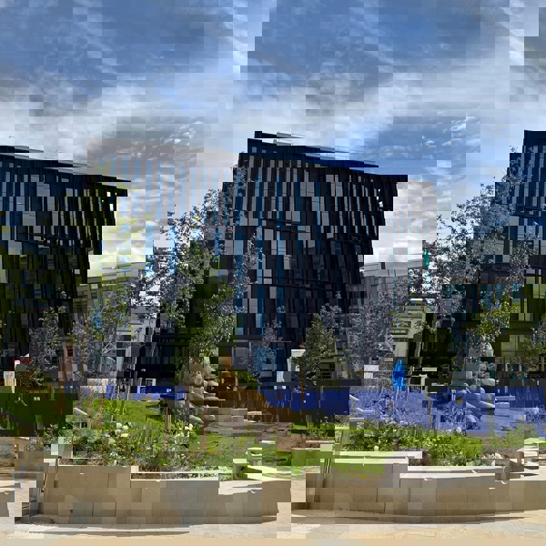 A modern black glass building with vertical panels stands behind a landscaped area featuring young trees, green grass, and large decorative rocks. Blue construction fencing surrounds part of the site, and the sky above is bright with scattered clouds and contrails.