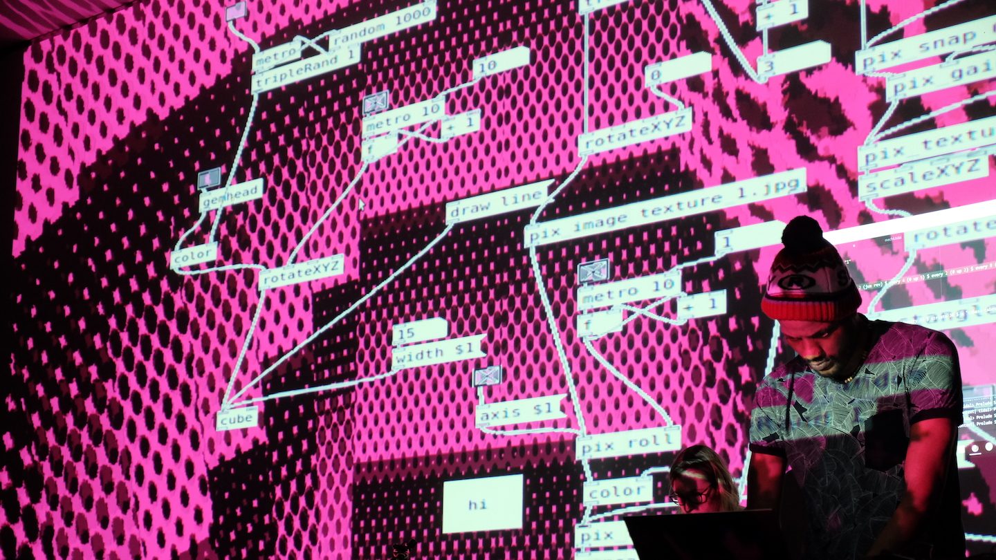 Person standing in front of a large projection displaying a visual programming interface with connected nodes and text blocks. The background is bright pink with black halftone patterns, and the projected text includes terms like ‘rotateXYZ,’ ‘pix image texture,’ and ‘metro 10.’