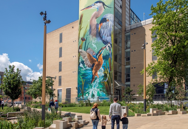 A large mural of two herons and a kingfisher in a vibrant wetland scene painted on the side of a tall modern building. In the foreground, a landscaped urban park with trees, benches, and pathways is visible, where several people, including a family with two children, are walking and enjoying the space under a bright blue sky with scattered clouds.