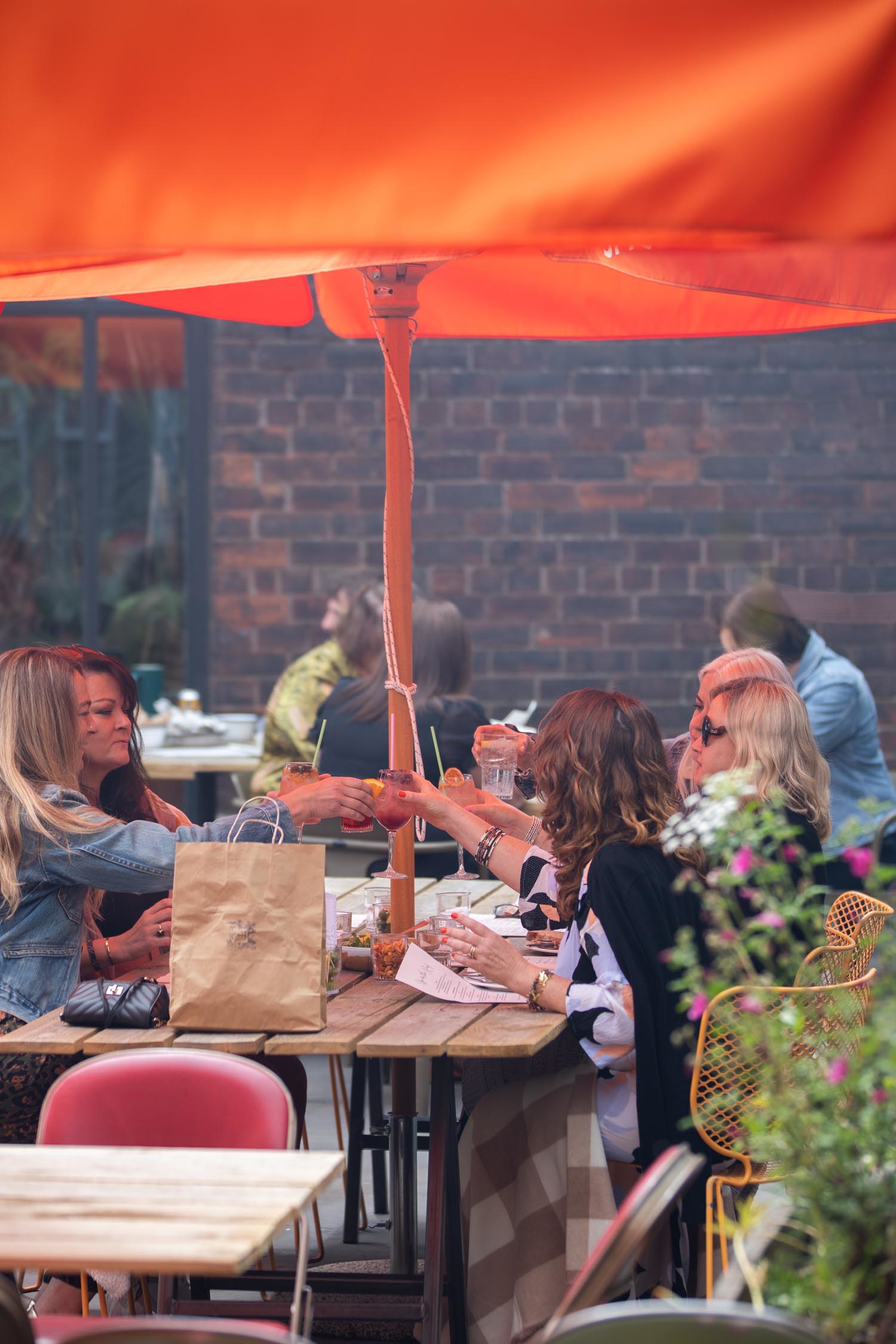 Group of people seated at an outdoor café table under a large orange umbrella, clinking glasses in a toast. The table has drinks, food, and a paper bag, with brick walls and other diners visible in the background.