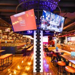Vibrant bar interior with wooden tables, orange seating, and large screens showing sports.
