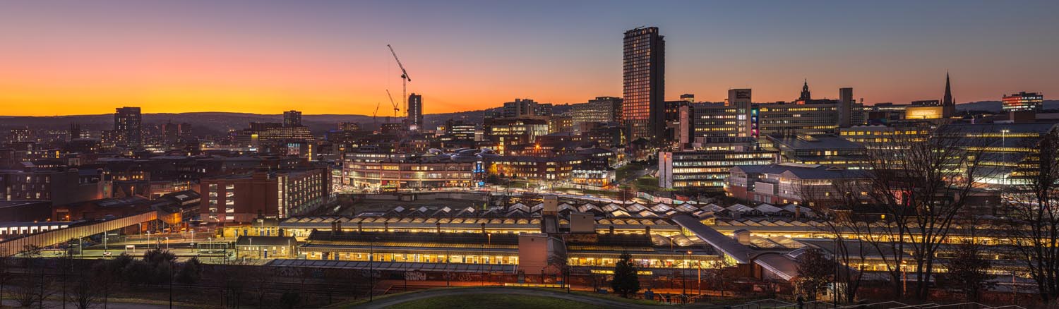 Panoramic view of a city skyline at sunset, with a vibrant orange and purple sky in the background. The scene features a mix of modern and historic buildings, including a tall tower and several mid-rise structures with illuminated windows. A construction crane is visible among the buildings, and a large train station with a glass roof occupies the foreground. Bare trees frame the lower edge of the image.
