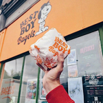 A hand holds a wrapped bagel from Best Boy Bagels, whose sign can be seen in the background.