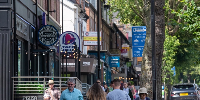 A row restaurants on a tree lined street. There are people walking up and down the pavement.