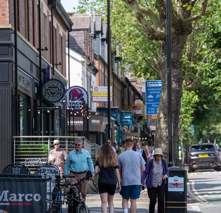 A row restaurants on a tree lined street. There are people walking up and down the pavement.