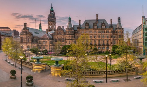 Panoramic view of Sheffield Town Hall and Peace Gardens at sunset. The historic stone building features ornate architecture with towers and a clock. In the foreground, landscaped gardens include fountains, trees, and paved walkways, surrounded by modern and traditional buildings.