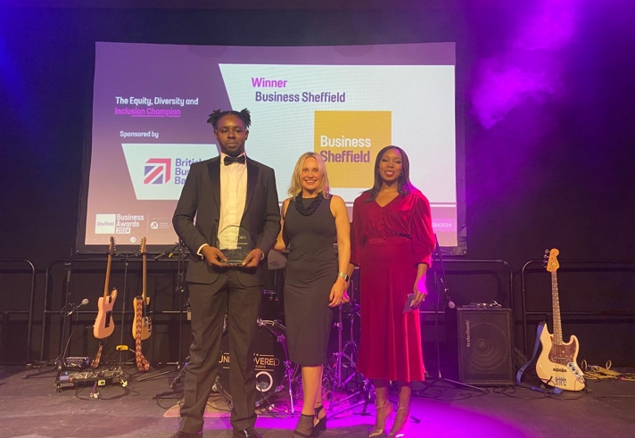 A group of people dressed in formal attire posing together at an indoor event with blue and purple stage lighting. Behind them is a stage with musical instruments and a large screen displaying “Sheffield Business Awards 2023” along with sponsor logos. One person in the center is holding an award, and another on the right is holding a framed certificate. The setting suggests a celebratory awards ceremony.