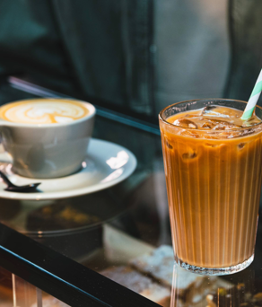 A close-up of two drinks on a glass counter in a coffee shop: a grey ceramic cup of latte with foam art on a saucer and spoon, and a tall glass of iced coffee with a green-and-white striped paper straw. Behind the drinks, part of a person’s jacket is visible, and jars of biscuits can be seen in the background.