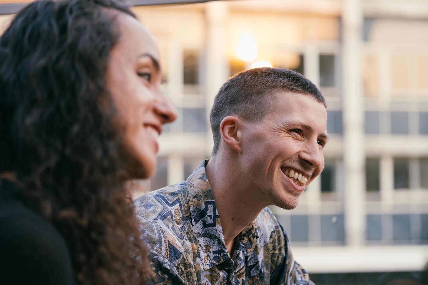 Two people sitting outdoors in front of a modern building with large windows and a blurred background. One person has long curly hair, and the other has short hair and is wearing a patterned shirt with geometric shapes in muted tones. Warm lighting suggests an evening or sunset setting.