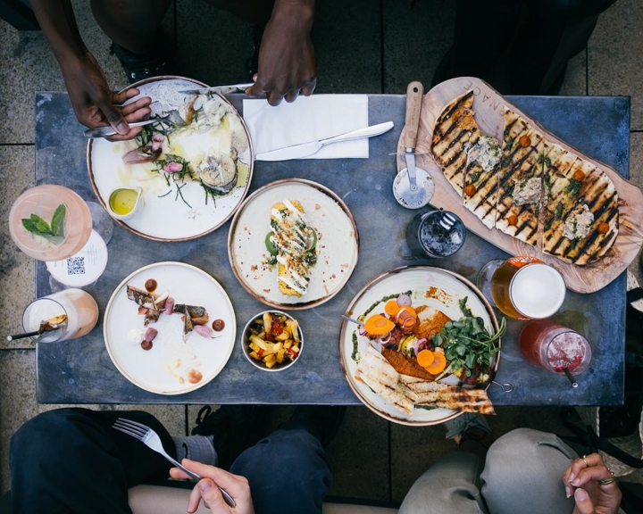 An overhead shot of a table in a restaurant filled with plates of food and drinks. There are people, mostly out of shot, enjoying the meal.