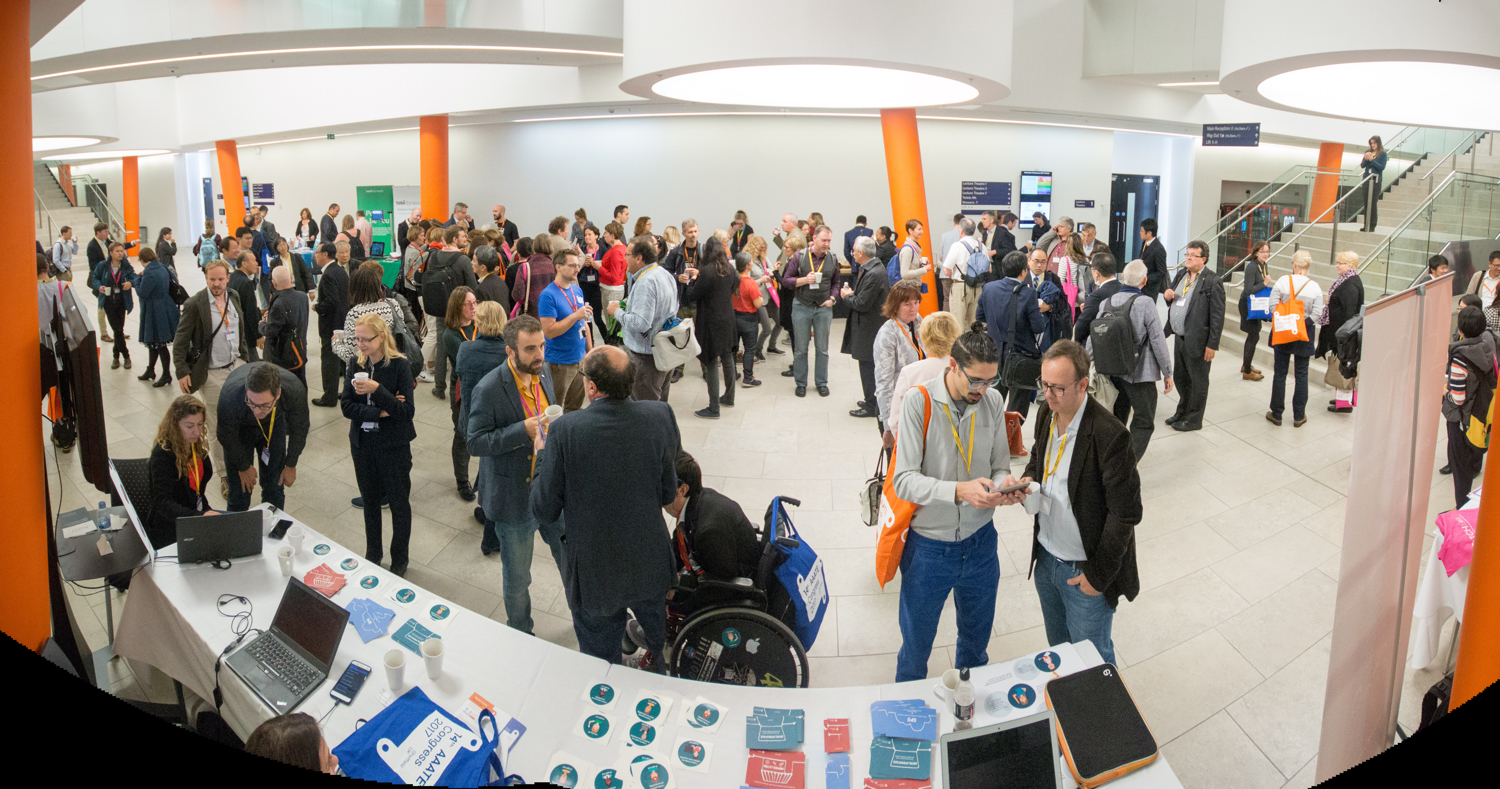Large group of people gathered in a bright, modern conference venue with white walls and orange pillars. Attendees are standing and conversing in the open space near registration tables displaying laptops, printed materials, and event badges. A staircase and signage are visible in the background.