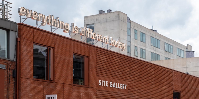Exterior of the red brick SITE Gallery building with the gallery name displayed on the wall and an 'everything is different today' illuminated sign and cloudy sky above.