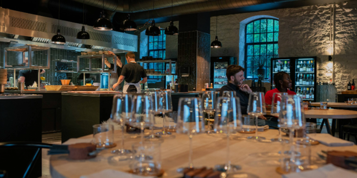 A modern, stylish restaurant interior featuring an open kitchen with a chef at work. In the foreground, a round wooden table is set with multiple empty wine glasses, folded napkins, and cutlery. The space has exposed stone walls, large arched windows, and shelves stocked with bottles. Black pendant lights hang from the ceiling, and the atmosphere is warm and contemporary.