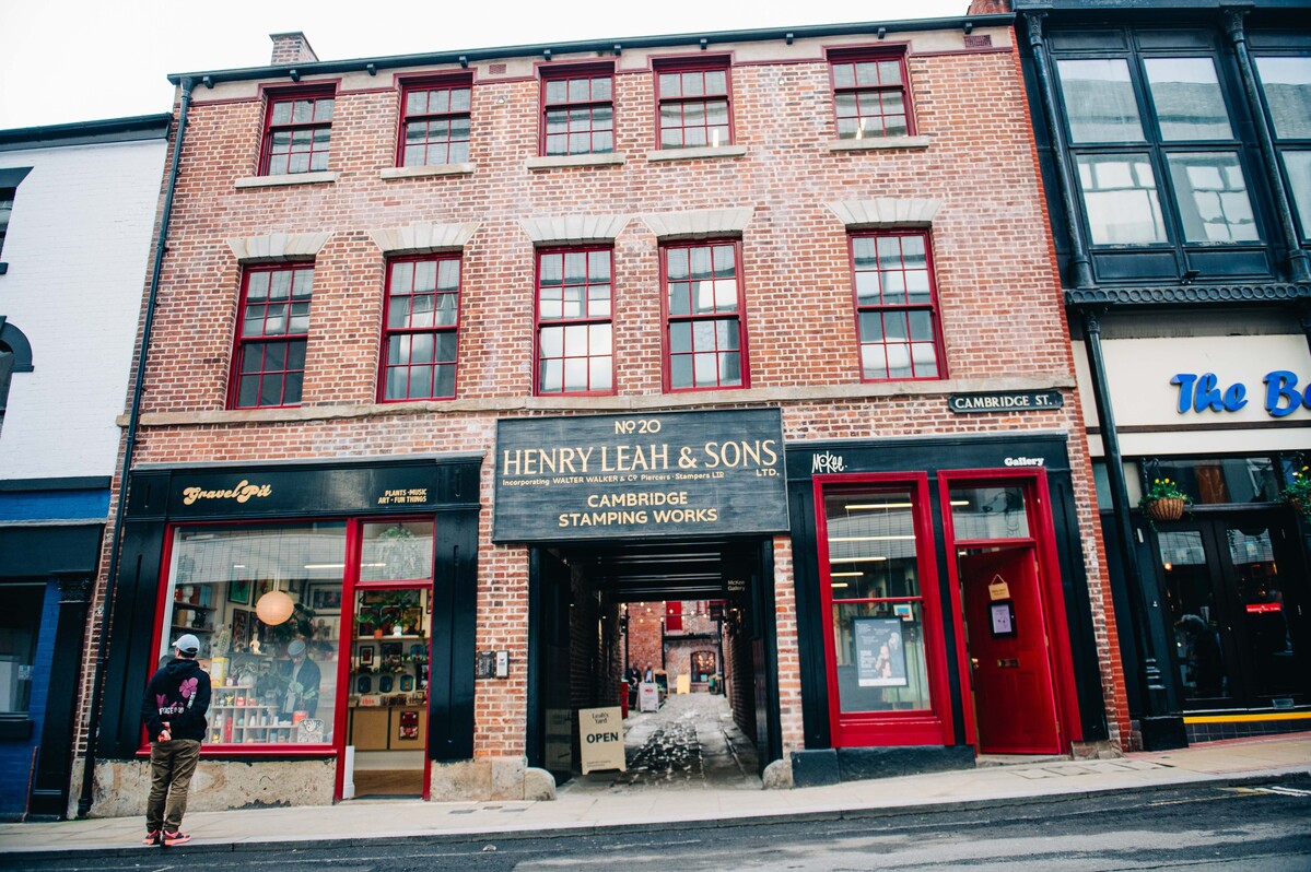 Exterior view of the historic red‑brick building at Leah’s Yard, featuring three floors of arched windows with red frames. The central entrance displays a large sign reading ‘Henry Leah & Sons Ltd – Cambridge Stamping Works’, with a cobbled passageway leading through to the courtyard beyond. Independent shopfronts with red doors and window displays sit on either side of the entrance.