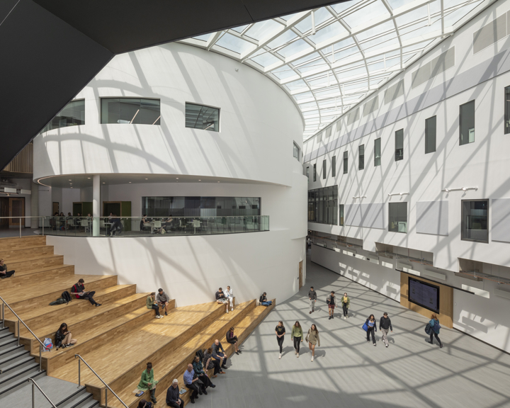 Spacious modern atrium with a curved white wall and glass ceiling allowing natural light to stream in. Wooden tiered seating occupies one side, where people are sitting and talking, while others walk across the open floor. The design features clean lines, large windows, and a bright, airy atmosphere.