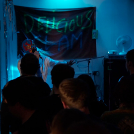 A performer wearing a bright, colorful mask and white outfit stands in front of a banner with neon-style text that reads “DELICIOUS CLAM.” The person is holding a microphone and gesturing toward the audience in a dimly lit room illuminated by blue lighting. Several people are gathered in the foreground, watching the performance, and metallic streamers hang along the wall to the left.