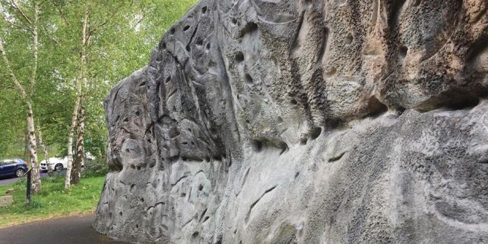 Outdoor climbing wall with various handholds and textures. It is located beside a paved path, surrounded by grass and trees, with parked cars visible in the background.