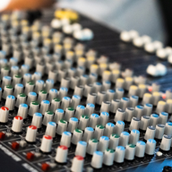 Close-up view of an audio mixing console with numerous knobs and sliders arranged in rows, featuring different coloured caps for channel control.