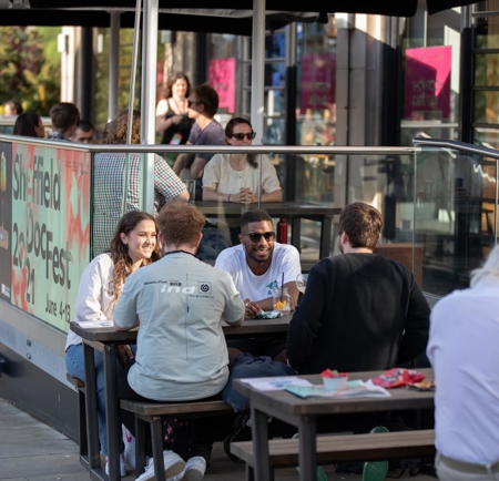 Outdoor seating area of a café or restaurant with several people sitting at wooden tables, eating and drinking. The space is enclosed by glass panels with colorful signage promoting “Sheffield DocFest” and event dates. String lights and umbrellas provide shade, and the background shows more tables and people inside the establishment.