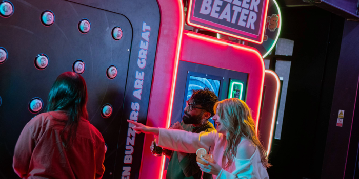 Three people playing an interactive arcade game called “Buzzer Beater” in a neon-lit venue. The game features a large vertical panel with multiple round buzzers and a digital score display reading “0550.” Bright signage on the machine includes phrases like “Green buzzers are great.” One person is actively playing, while another points at the panel, holding a drink. The setting is vibrant with colourful lights and a lively atmosphere.
