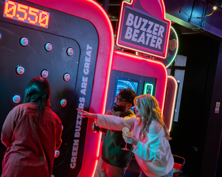 Three people playing an interactive arcade game called “Buzzer Beater” in a neon-lit venue. The game features a large vertical panel with multiple round buzzers and a digital score display reading “0550.” Bright signage on the machine includes phrases like “Green buzzers are great.” One person is actively playing, while another points at the panel, holding a drink. The setting is vibrant with colourful lights and a lively atmosphere.