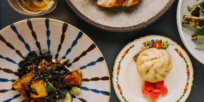 Top-down view of a table with assorted dishes, including a plate of roasted vegetables with greens, a decorative plate holding a steamed bun and pickled radishes, a stone bowl with grilled meat topped with a lemon wedge, and a glass of golden-coloured drink.