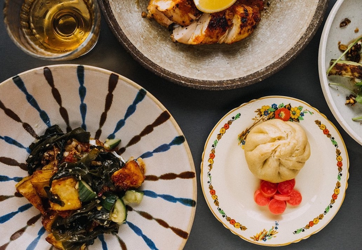 Top-down view of a table with assorted dishes, including a plate of roasted vegetables with greens, a decorative plate holding a steamed bun and pickled radishes, a stone bowl with grilled meat topped with a lemon wedge, and a glass of golden-coloured drink.