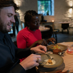 Two people seated at a wooden table in a modern restaurant, enjoying a plated dish. The table holds ceramic plates with artfully presented food, cutlery, and glasses with drinks. The background shows softly lit stone walls, other tables, and a large window, creating a warm and contemporary dining atmosphere.