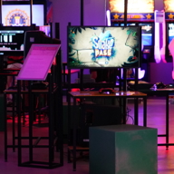 Gaming area with a monitor displaying the game ‘Snake Pass’ on a central table, surrounded by green cube seats. In the background, several arcade machines with bright screens and colourful designs are visible, creating a vibrant atmosphere.