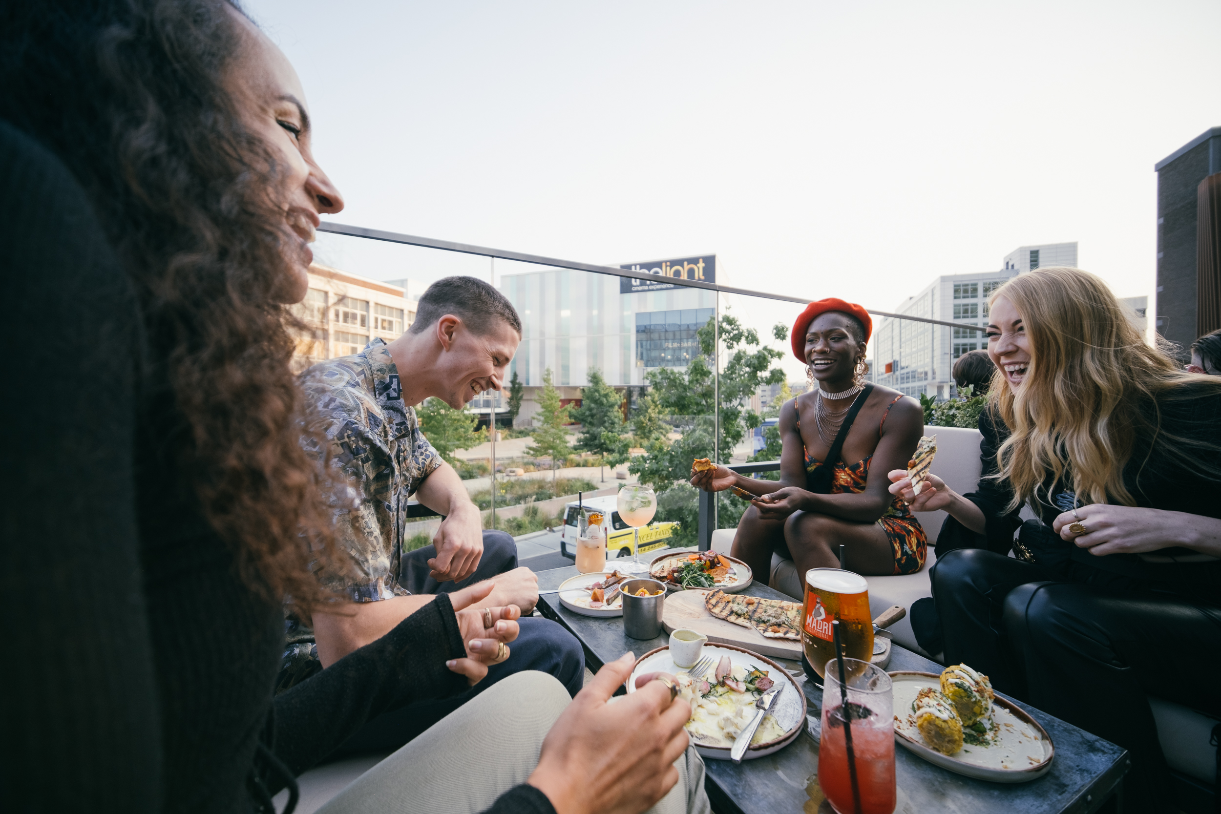Group of people sitting at an outdoor rooftop table with plates of food and drinks. The table has tacos, sandwiches, and cocktails, and the background shows modern buildings and greenery under a clear sky.