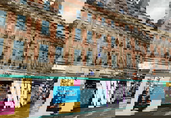 Street view of a historic building with a Radisson Blu hotel sign, featuring ornate windows and black wrought-iron balconies. In front of the building is a colorful construction hoarding with promotional graphics and text, including phrases like “The Northern Capital of Cool” and “WHEN.” The hoarding displays images of people indoors, event details, and branding elements. Scaffolding is visible along the lower part of the building, and the sky is overcast.