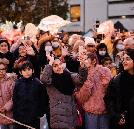 Crowd gathered at an outdoor event during dusk, with people standing behind a rope barrier and raising hands or holding phones. Illuminated lanterns and lighted decorations are visible in the background, along with buildings and trees.