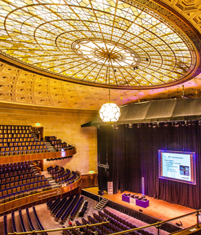 The Oval Hall at Sheffield City Hall. Grand auditorium with tiered seating and a large stage featuring a projection screen. The ornate ceiling has an intricate geometric design with a central stained-glass dome and a hanging light fixture. The stage is framed by black curtains, and the seating area includes multiple balconies with curved railings.