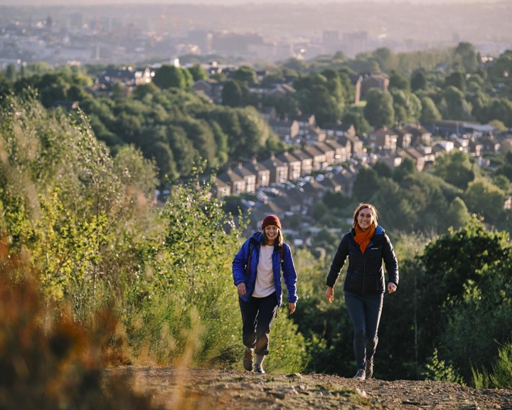 Two ramblers are walking up a hill. Down in the valley below them you can see rows of terrace houses and on the horizon you can see Sheffield city centre.