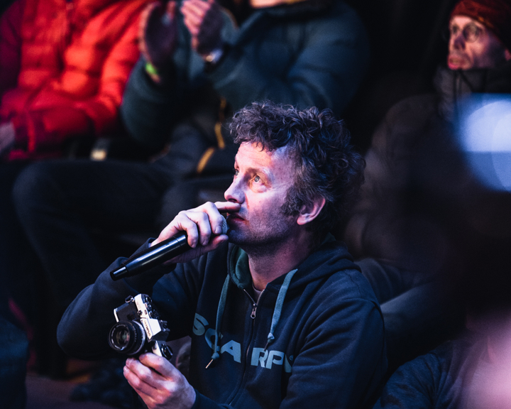 A man is sat in a group holding a microphone and a camera.