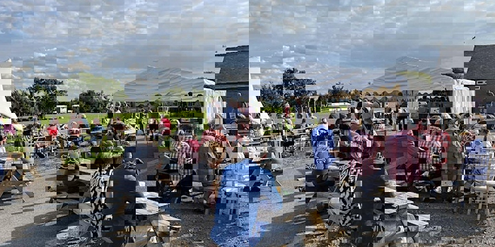 A sunny outdoor scene at a beer festival with groups of people sitting at wooden picnic benches on a paved area beside a large grassy field. A white marquee tent is set up in the background with people gathered around it. The sky is bright with scattered clouds.