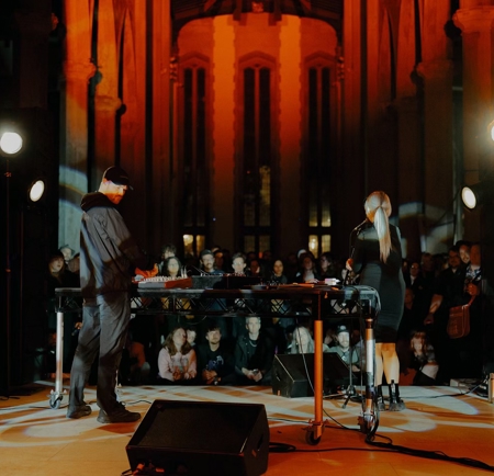 Two performers stand behind a DJ table in a grand hall with tall columns and arched windows, illuminated by warm orange lighting and spotlights. A seated audience watches in the background.