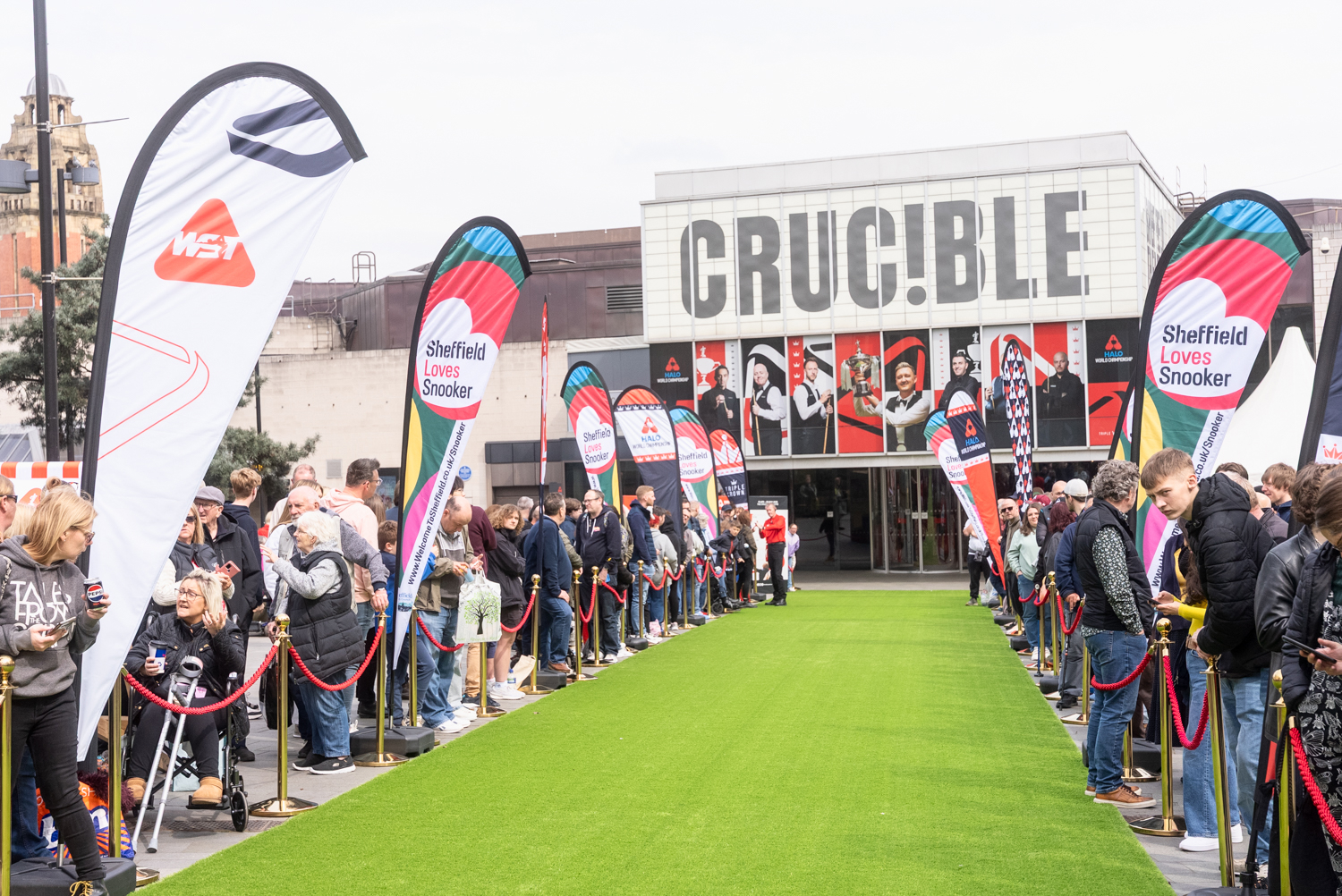 Crowds line a green carpet outside the Crucible Theatre for a Sheffield Loves Snooker event, with colourful flags and the theatre’s bold façade in the background.